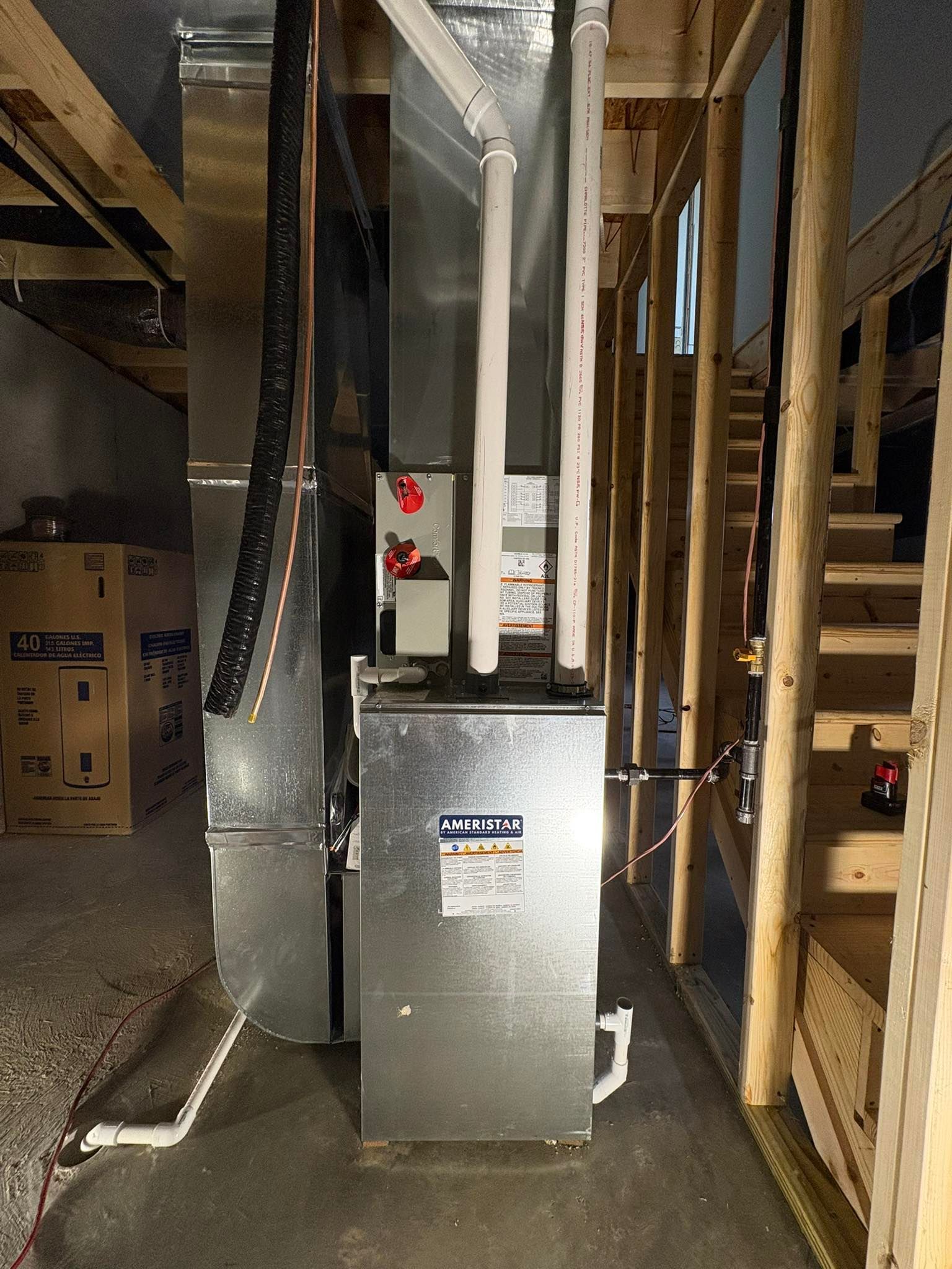 A metal furnace unit in an unfinished basement, with white PVC piping, next to wooden stair framing.