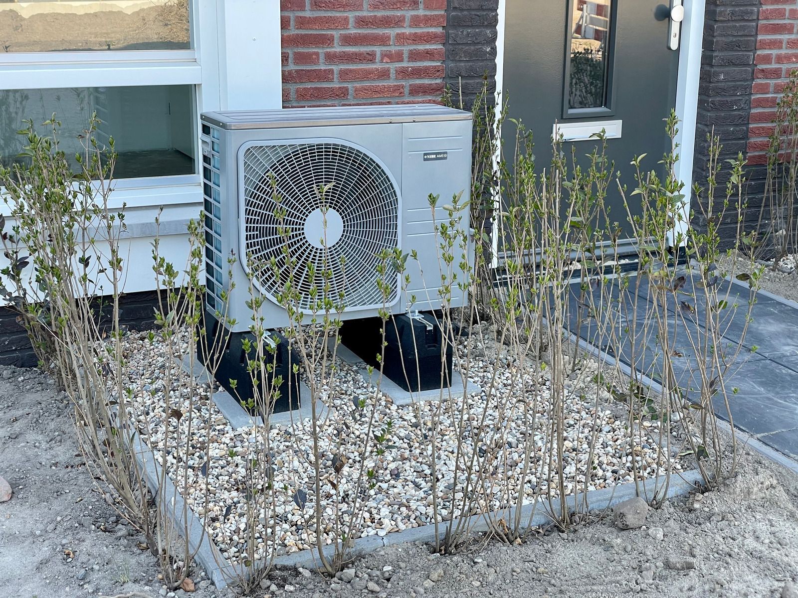 An outdoor heat pump unit sits on black stands atop a gravel bed surrounded by small hedges in front of a house.
