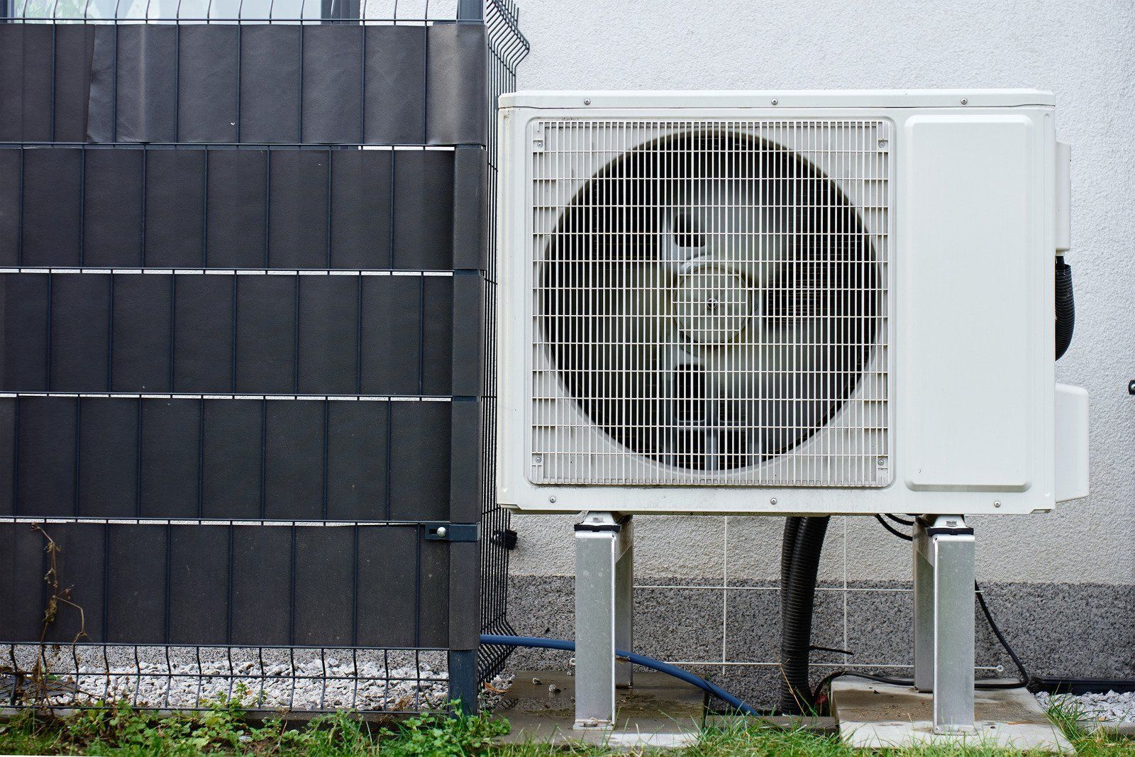 White heat pump outdoor unit mounted on metal legs beside a dark privacy fence and a building wall.