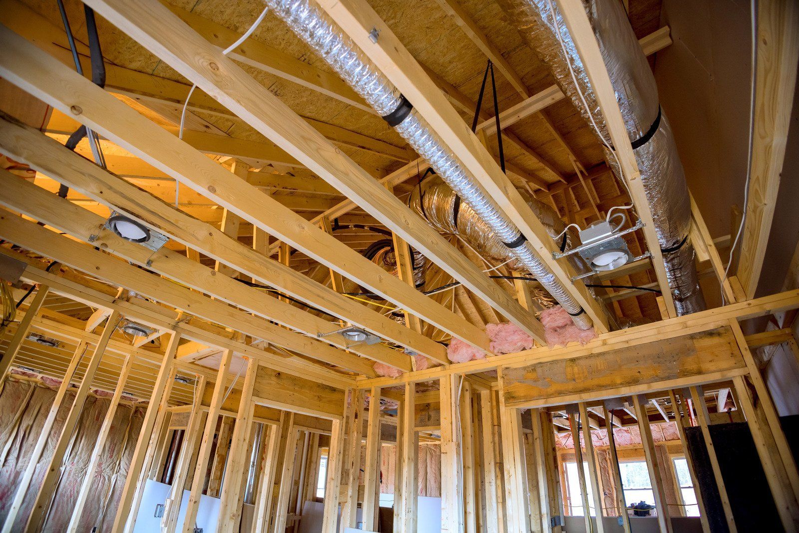 Wooden framing and rafters of a house under construction, with exposed HVAC ducting and recessed lighting fixtures.