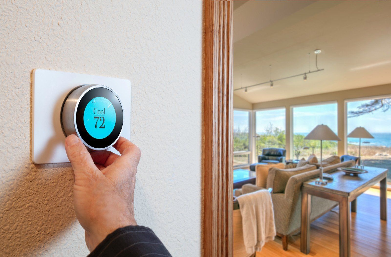A person adjusts a circular, blue-lit smart thermostat on a beige wall, overlooking a bright living room with ocean views.