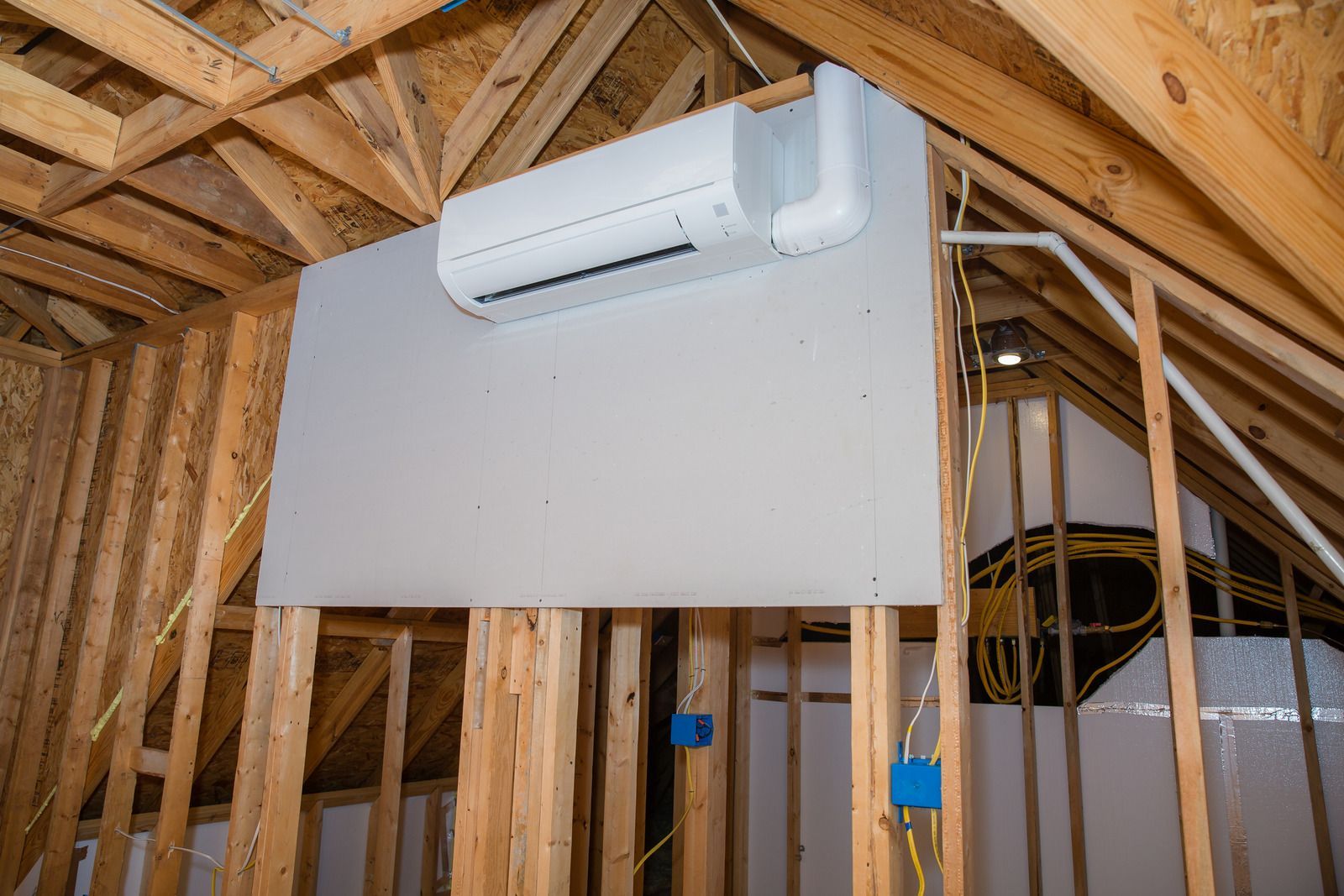 A mini-split air conditioning unit mounted on a drywall panel within the wooden frame of an unfinished attic.