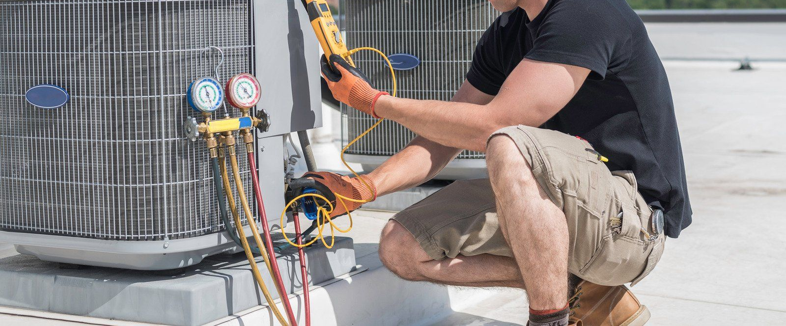 A technician in work clothes crouches on a rooftop, using gauges and a multimeter to service an HVAC condenser unit.