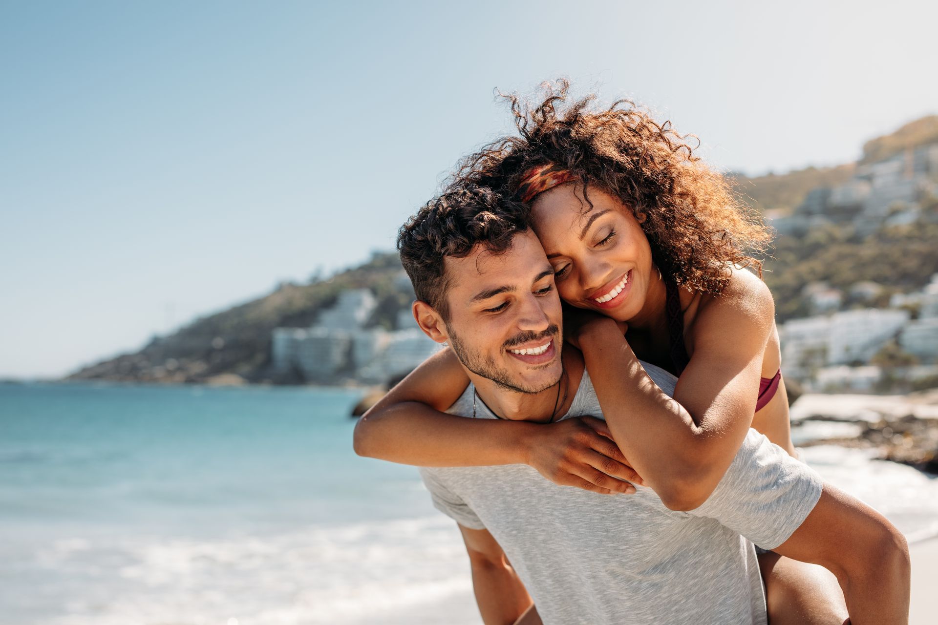 Man giving woman a piggyback ride on the beach, smiling. Ocean and mountains in the background.