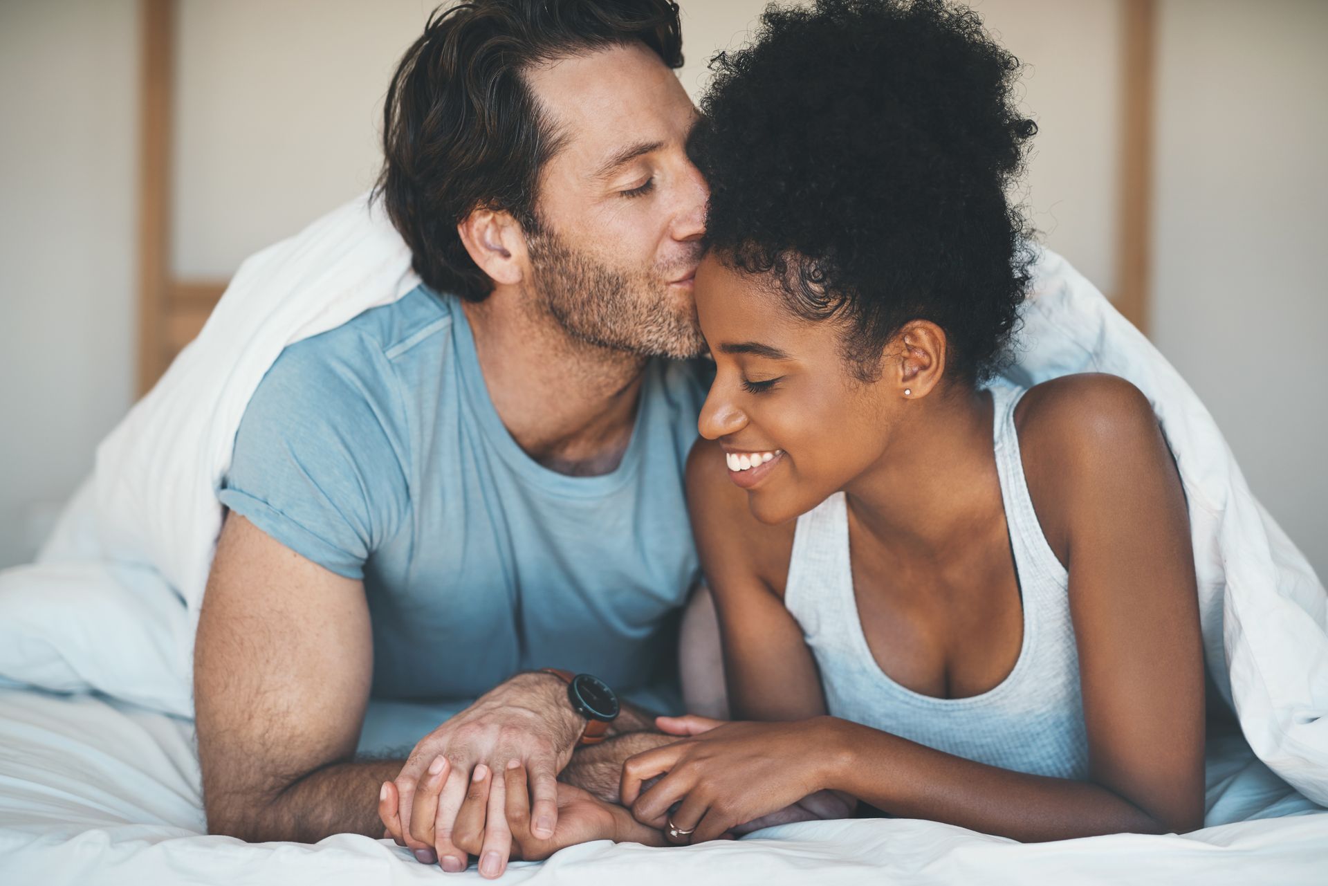 Man kissing a Black woman's forehead while they lie on a bed, smiling under a blanket.