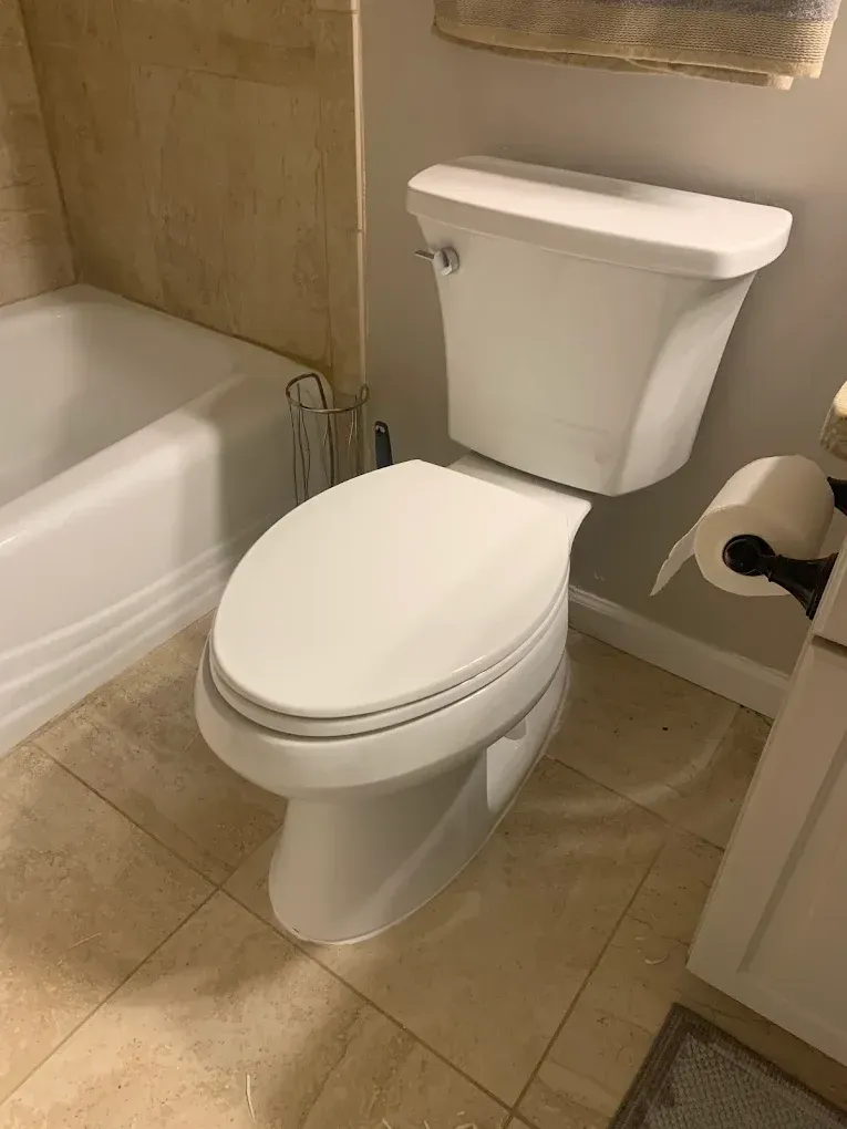 White toilet in a bathroom next to a bathtub. Light brown tile floor, neutral-colored walls.