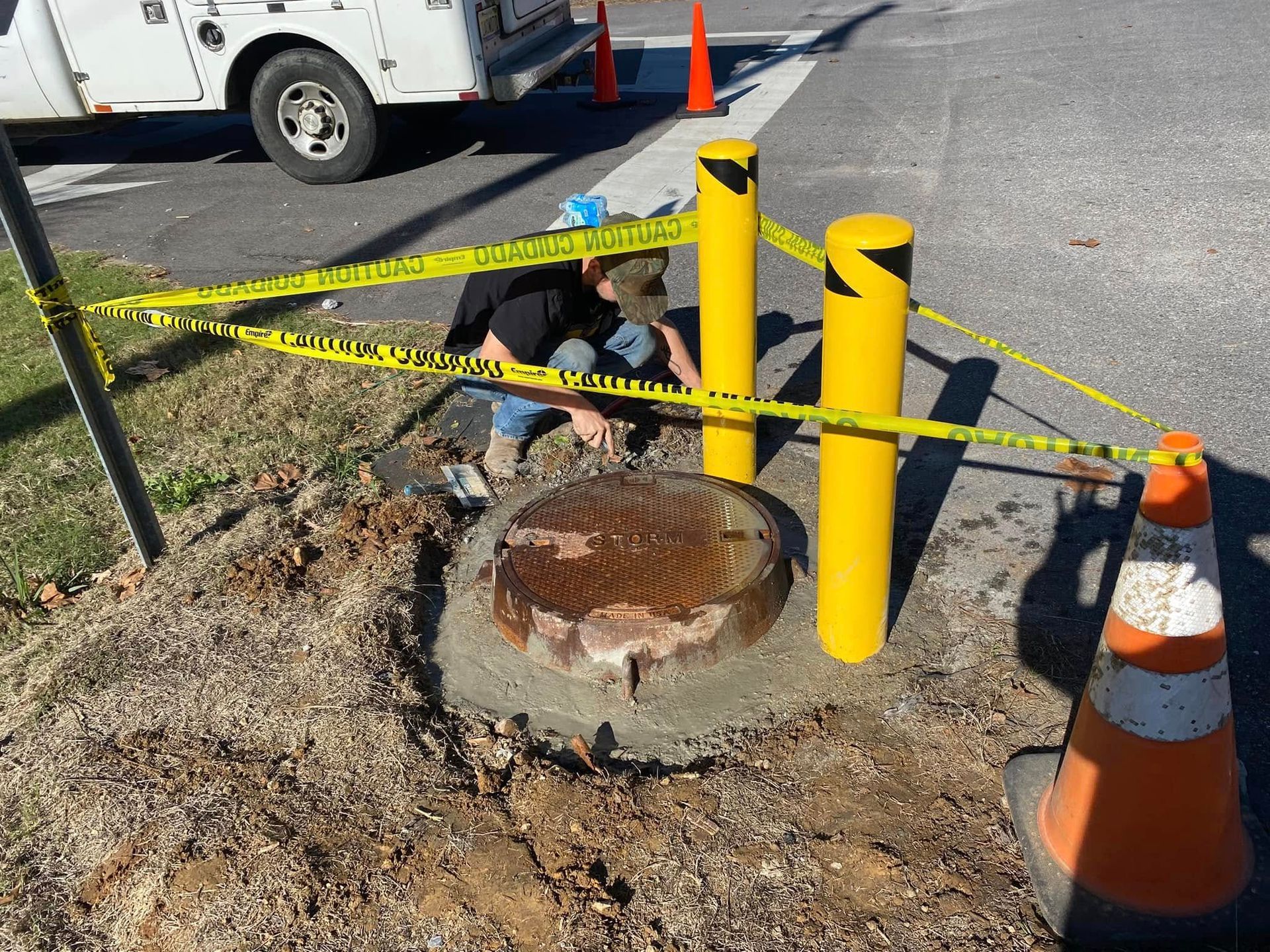 Man working near a manhole cover surrounded by yellow bollards, caution tape, and an orange traffic cone.