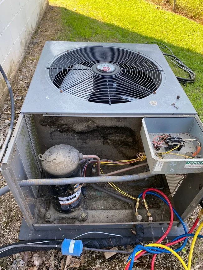 An open outdoor air conditioner unit with fan, compressor, and wiring. Grey and metal in a grassy yard.