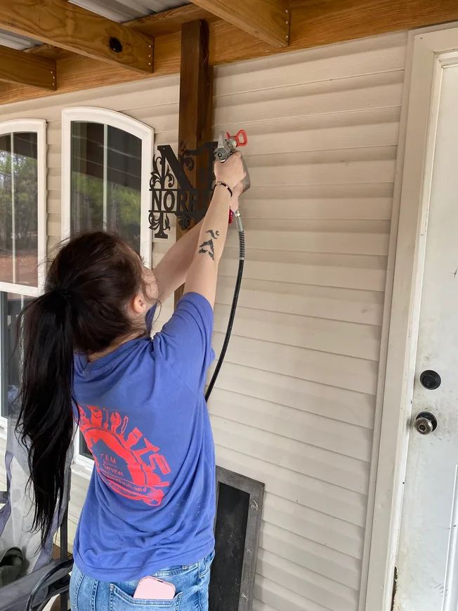 Woman spraying siding brown with a paint sprayer on a porch; exterior.