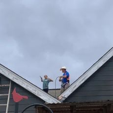 Two people on a gray rooftop, one gesturing while the other uses equipment; cloudy sky.