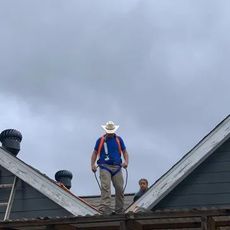 Man on a roof wearing a cowboy hat and safety harness, working with another person, cloudy sky.