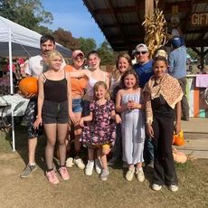 Group poses at an outdoor fall market: people of different ages, pumpkins, and a gift stand.