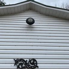 White siding on a building with a black outdoor light fixture. Decorative metalwork is below the light.