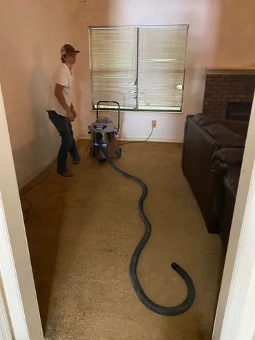 Man vacuuming carpet in a room. The carpet is beige, and the walls are white. There is a window and a fireplace.