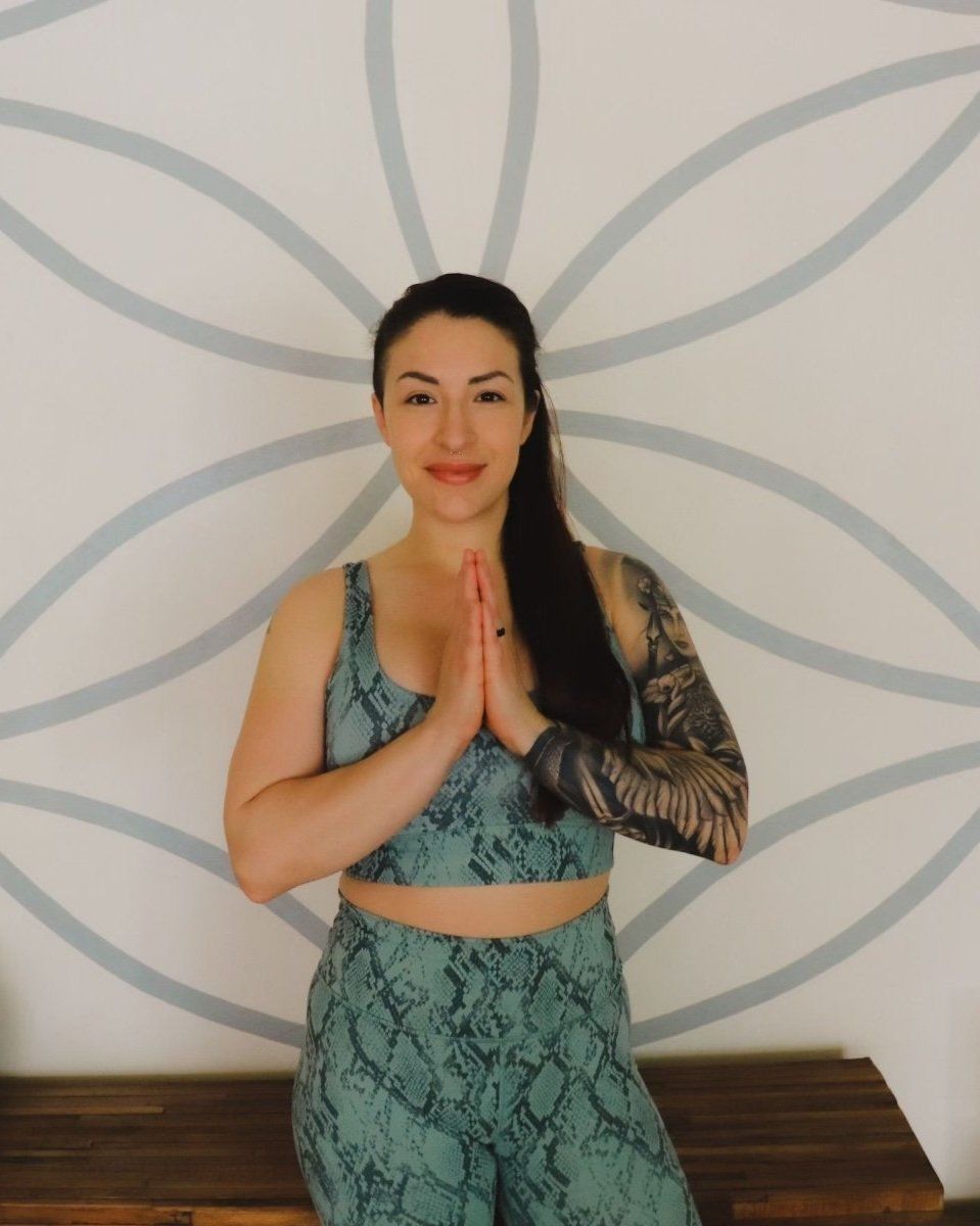 A woman with a tattoo on her arm is sitting on a bench with her hands folded in front of a wall.