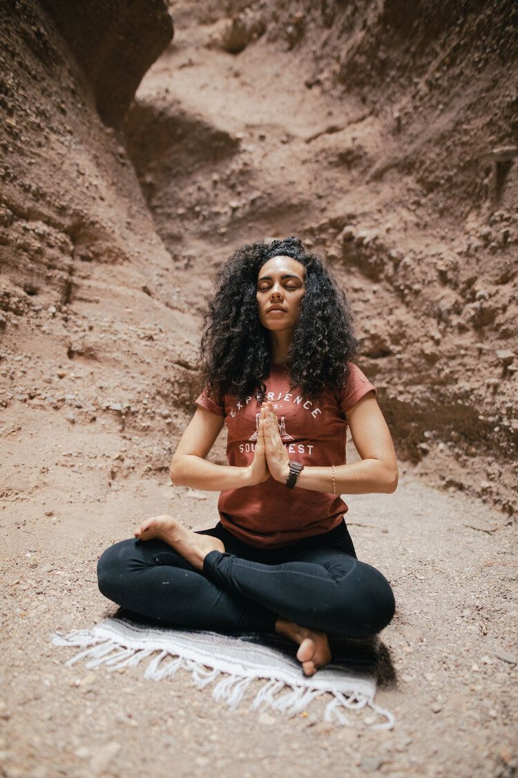 A woman is sitting in a lotus position on a yoga mat in the dirt.