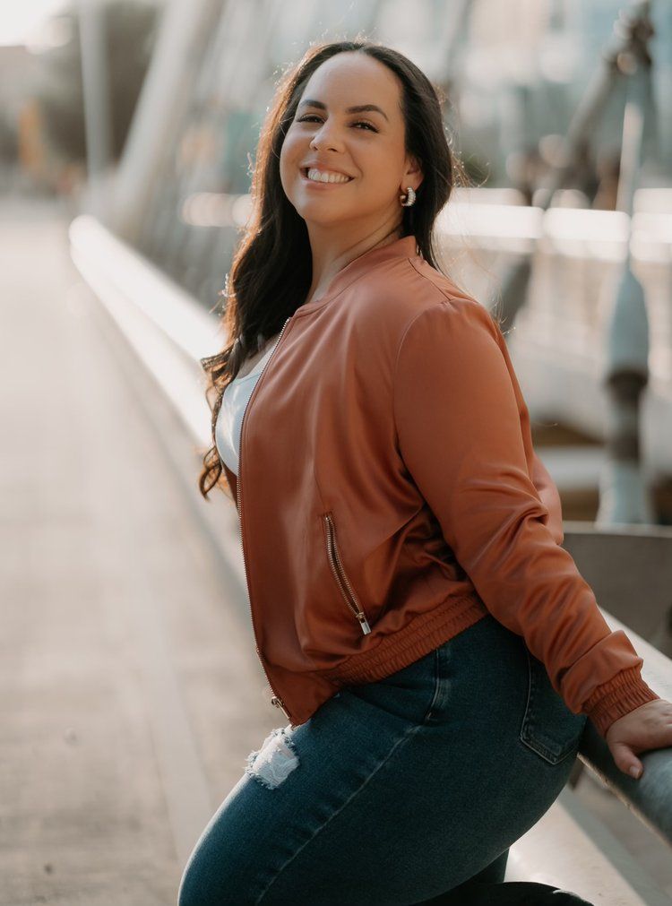 A woman in a brown jacket and blue jeans is sitting on a railing.