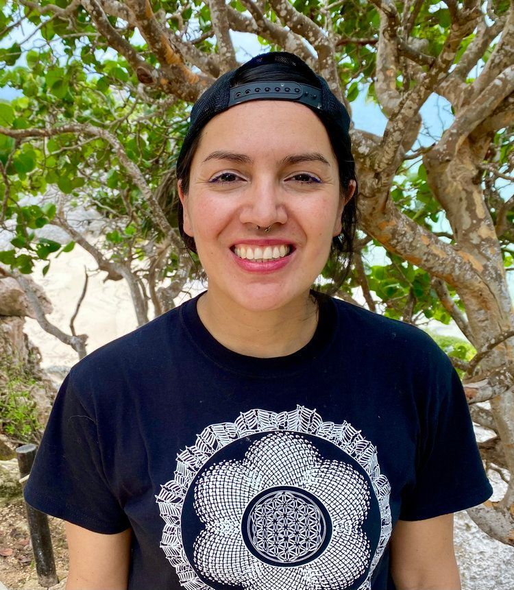A woman wearing a hat and a black t-shirt is smiling in front of a tree.