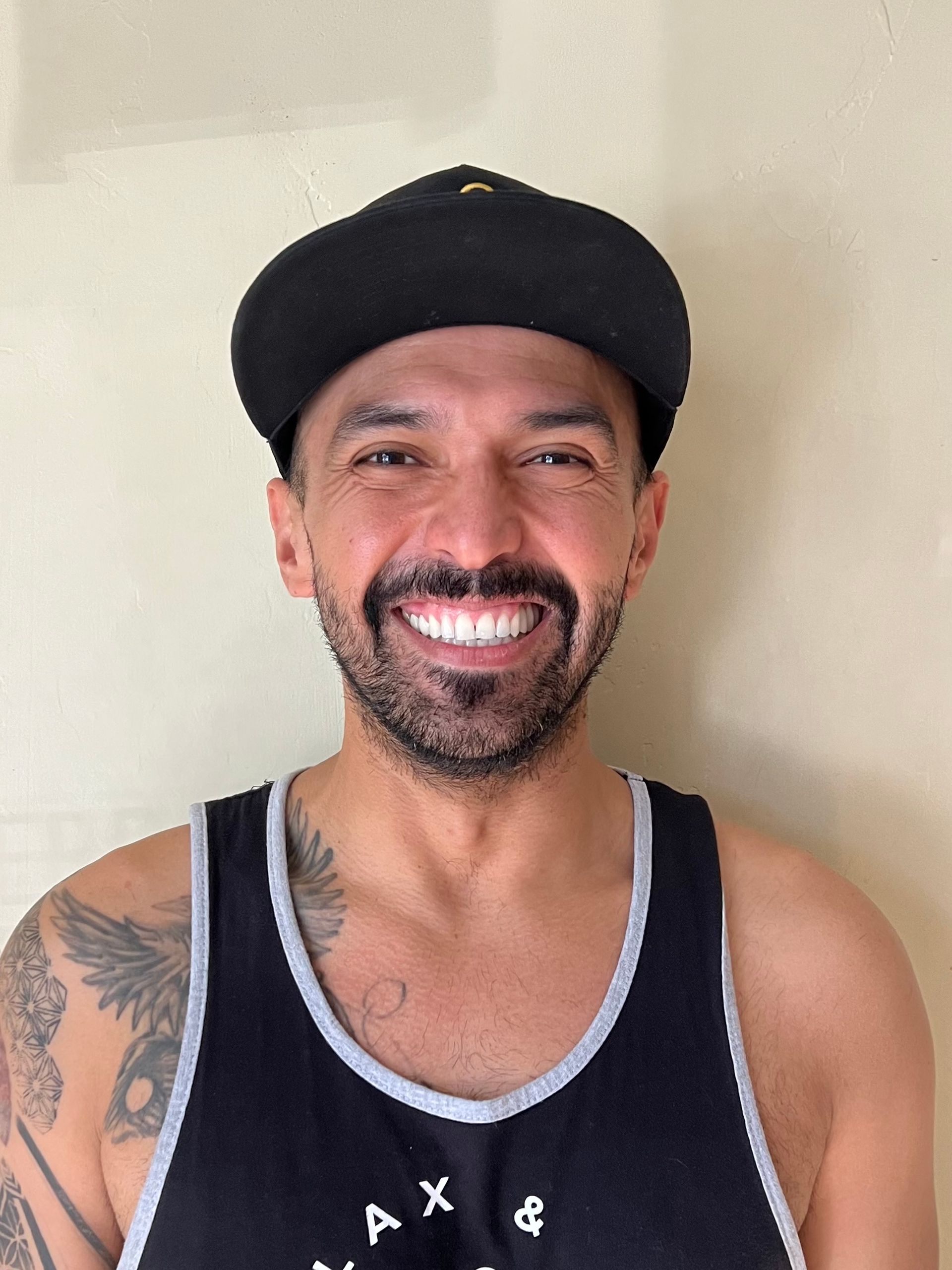 A man with a beard wearing a hat and a tank top is smiling.