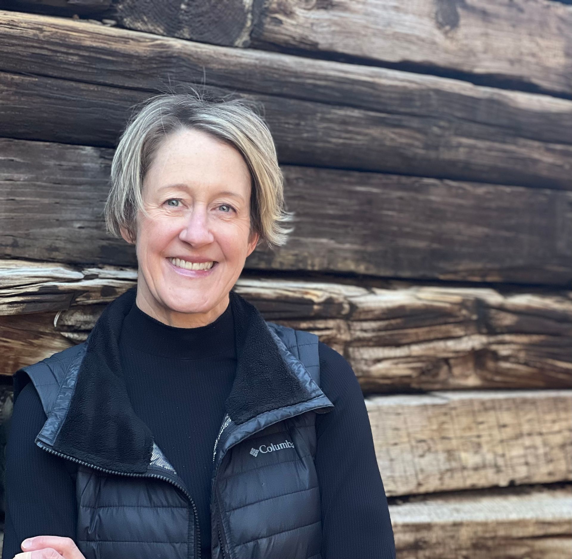 A woman in a black vest is smiling in front of a wooden wall.