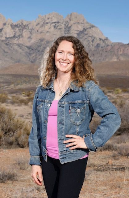 A woman in a denim jacket and a pink shirt is standing in front of a mountain.