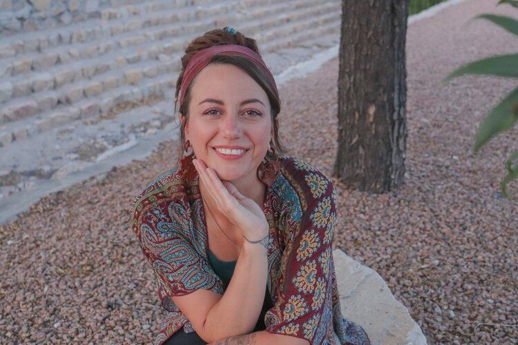 A woman is sitting on a rock with her hand on her chin and smiling.