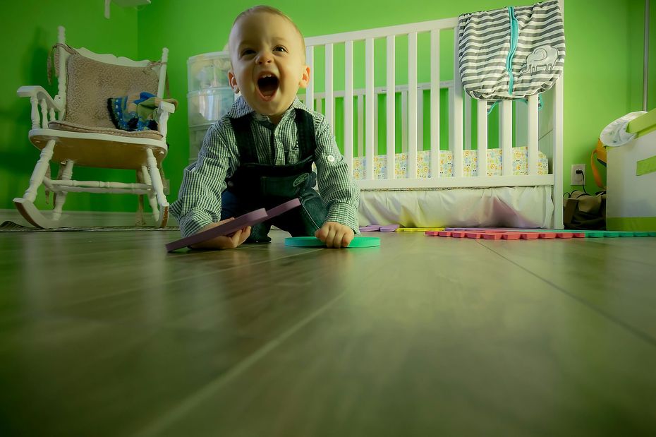 A delighted toddler kneels on a wood floor, laughing with mouth open, near toys. Green wall, white crib and rocker.