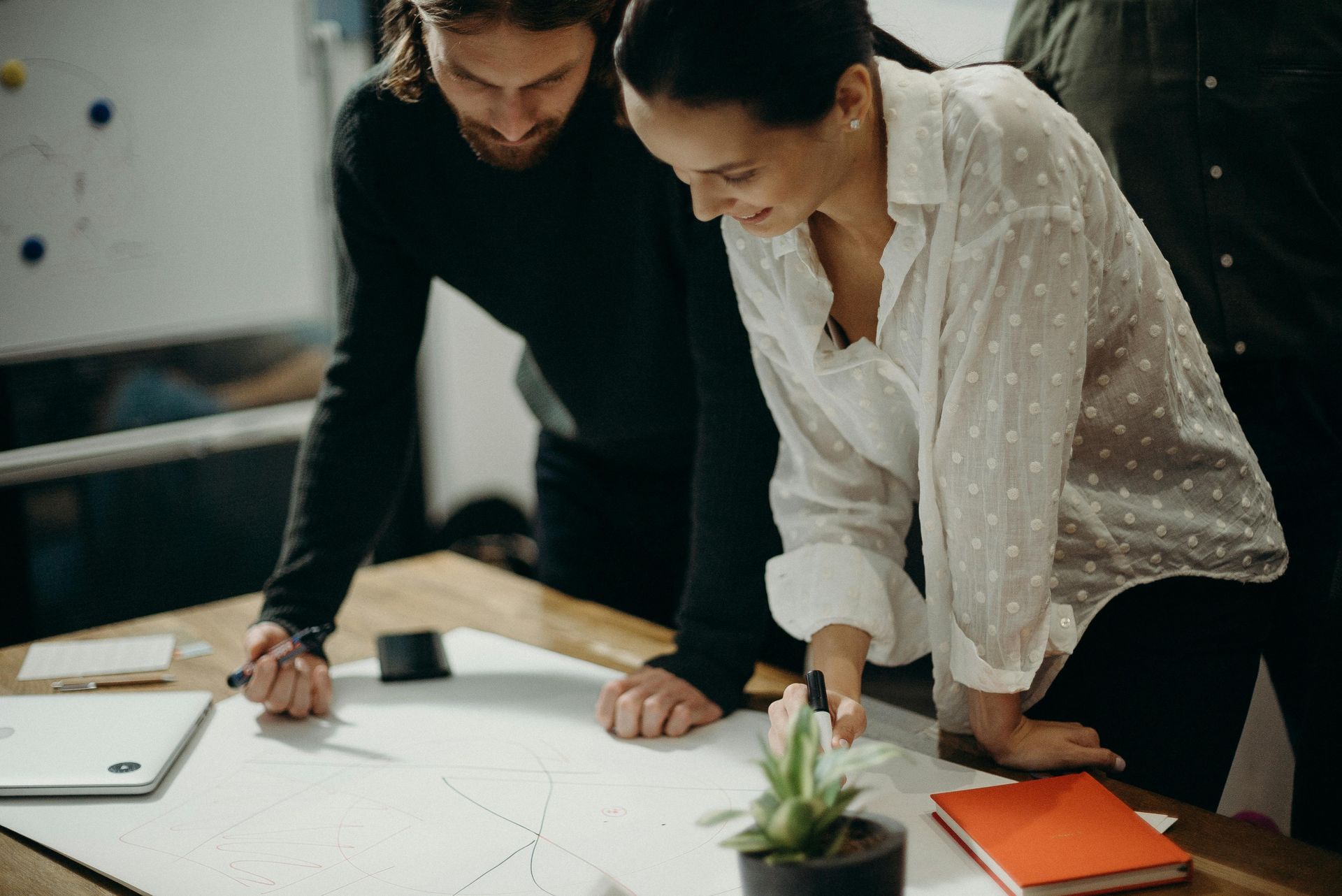 Two people lean over a table examining a document, a plant and a notebook are on the table.