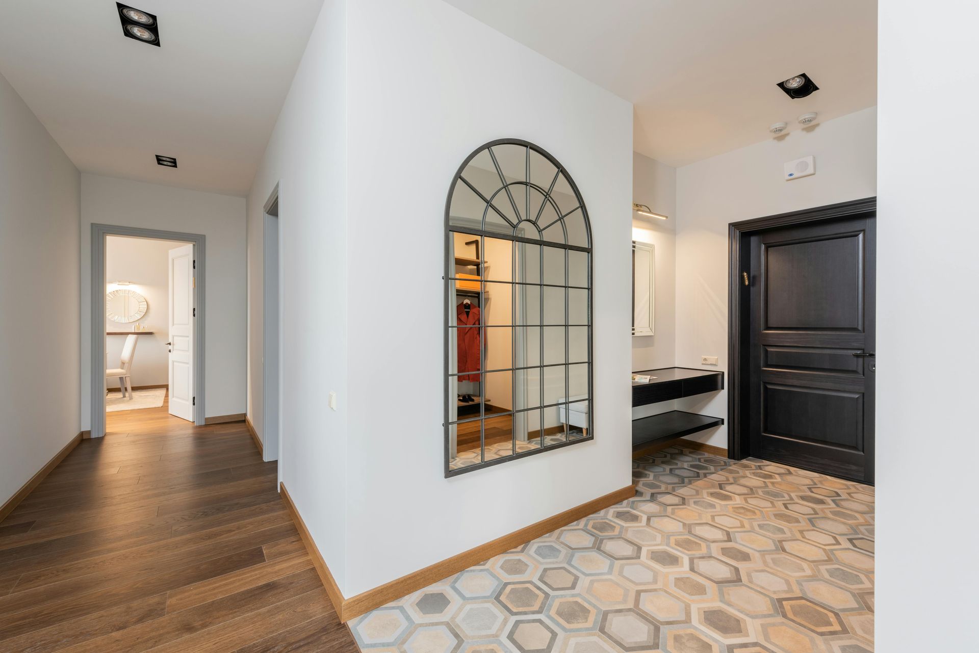 Hallway with a decorative arched mirror, dark wood floors, and patterned tile. A black door and white walls are present.