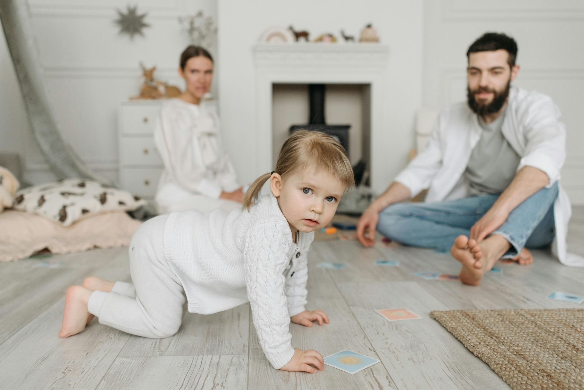 Girl crawling on the floor toward the camera, parents in background, white room.