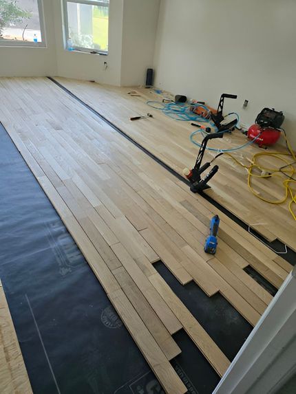 Installing hardwood flooring in a room. Boards laid, tools on floor: nailer, compressor, and power cords.