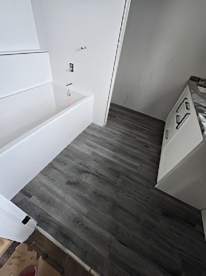 White bathroom with gray wood-look flooring. Tub, shower fixtures, and cabinet visible.
