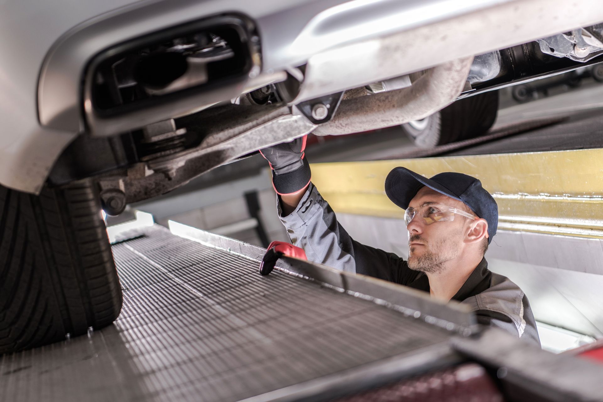 Mechanic in gloves examines a car's undercarriage on a lift. He wears glasses, a cap, and work clothes.