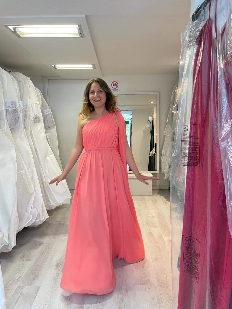 Una donna con un lungo abito rosa è in piedi in un negozio di abiti da sposa.