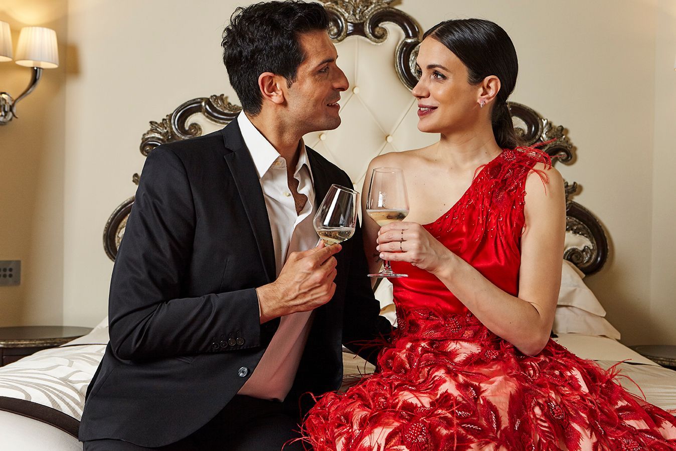 Un uomo e una donna sono seduti su un letto e bevono vino. La donna indossa un vestito rosso.