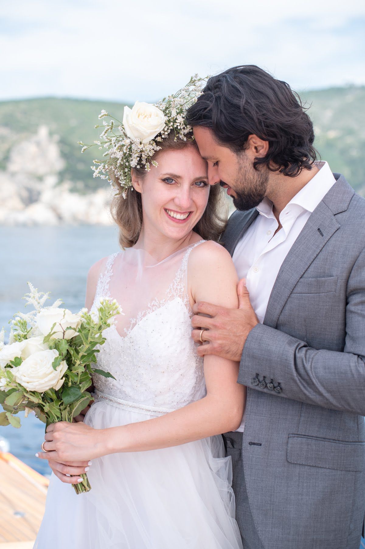 Una sposa e uno sposo posano per una foto di fronte all'oceano. La sposa indossa una corona di fiori.