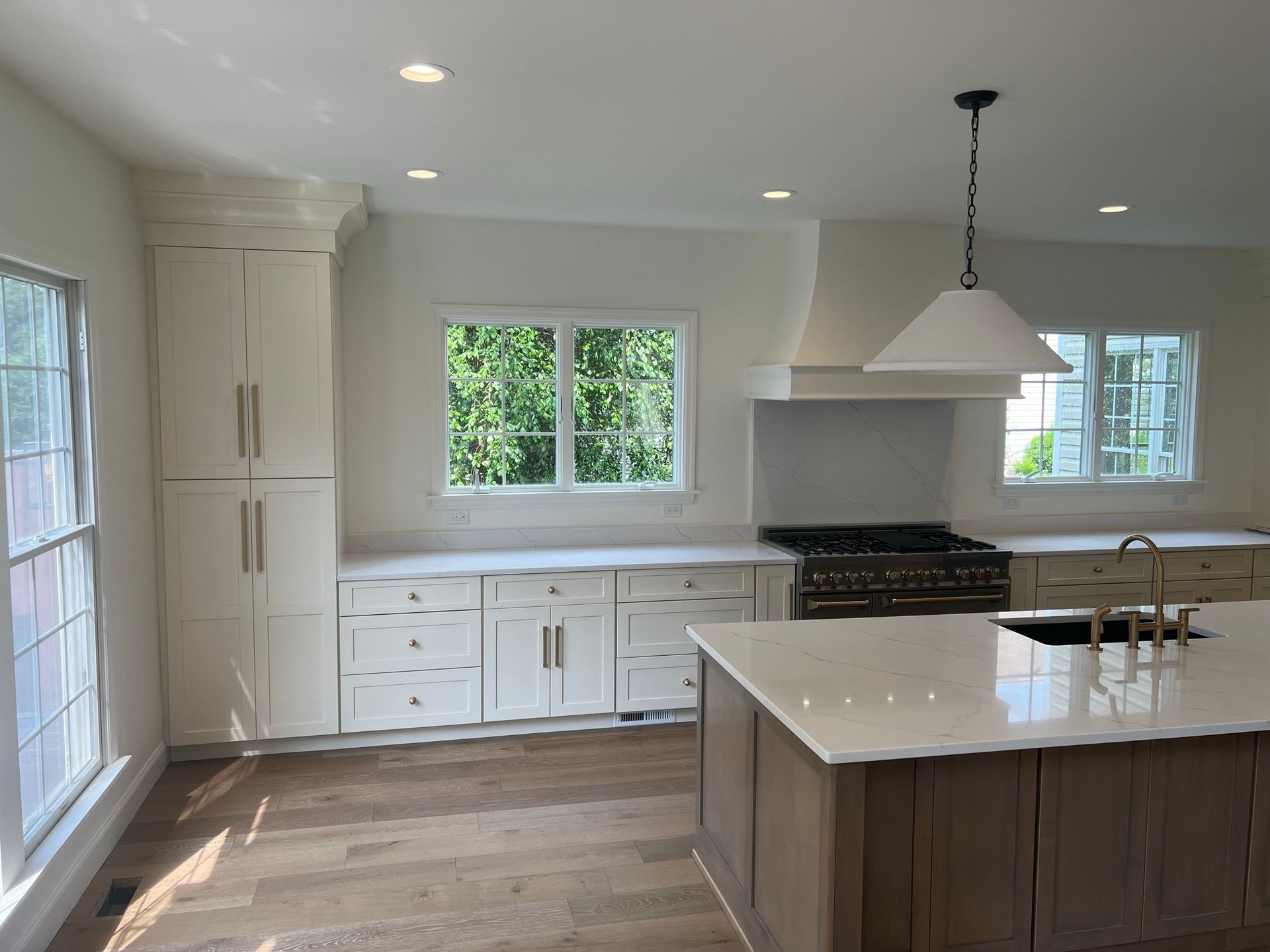 Modern white kitchen with island, wood floors, pendant light, and large windows.