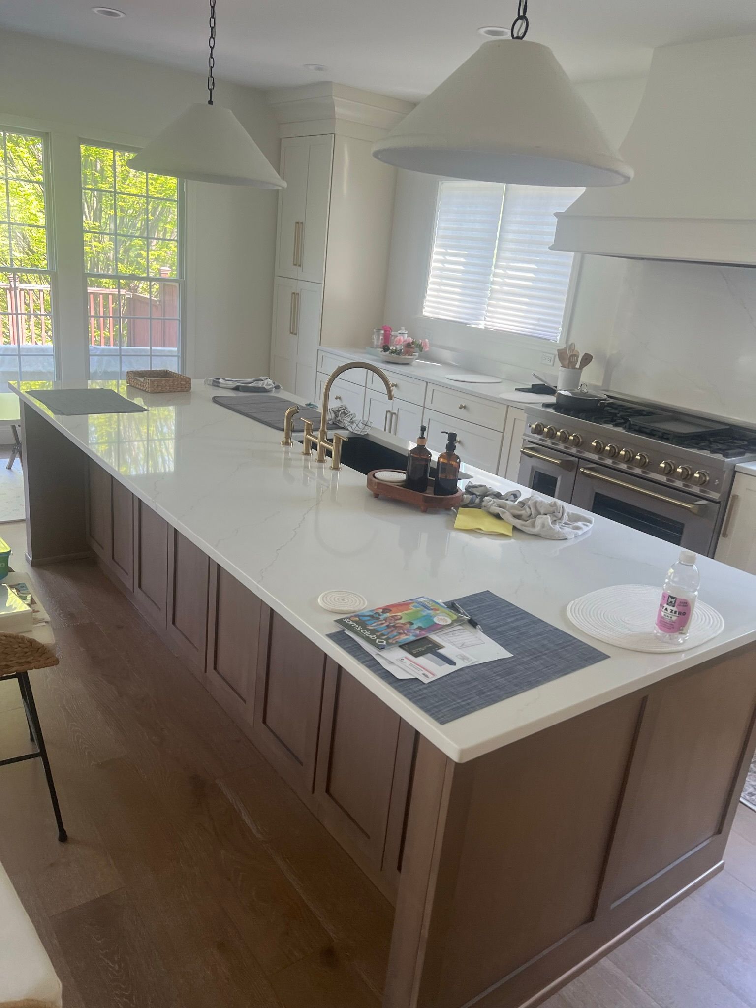 Bright kitchen with large island, white countertops, pendant lights, and stainless steel stove by windows.