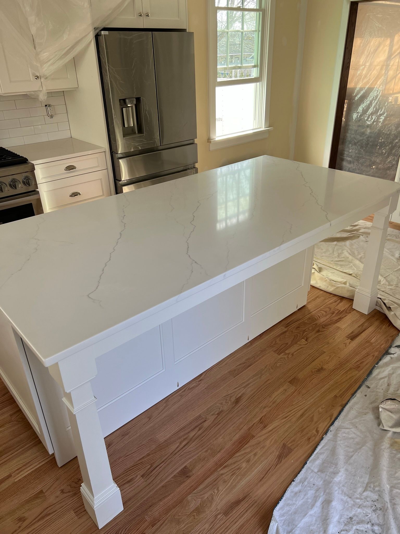 White kitchen island with glossy countertop, next to stainless fridge and window, over wood floor