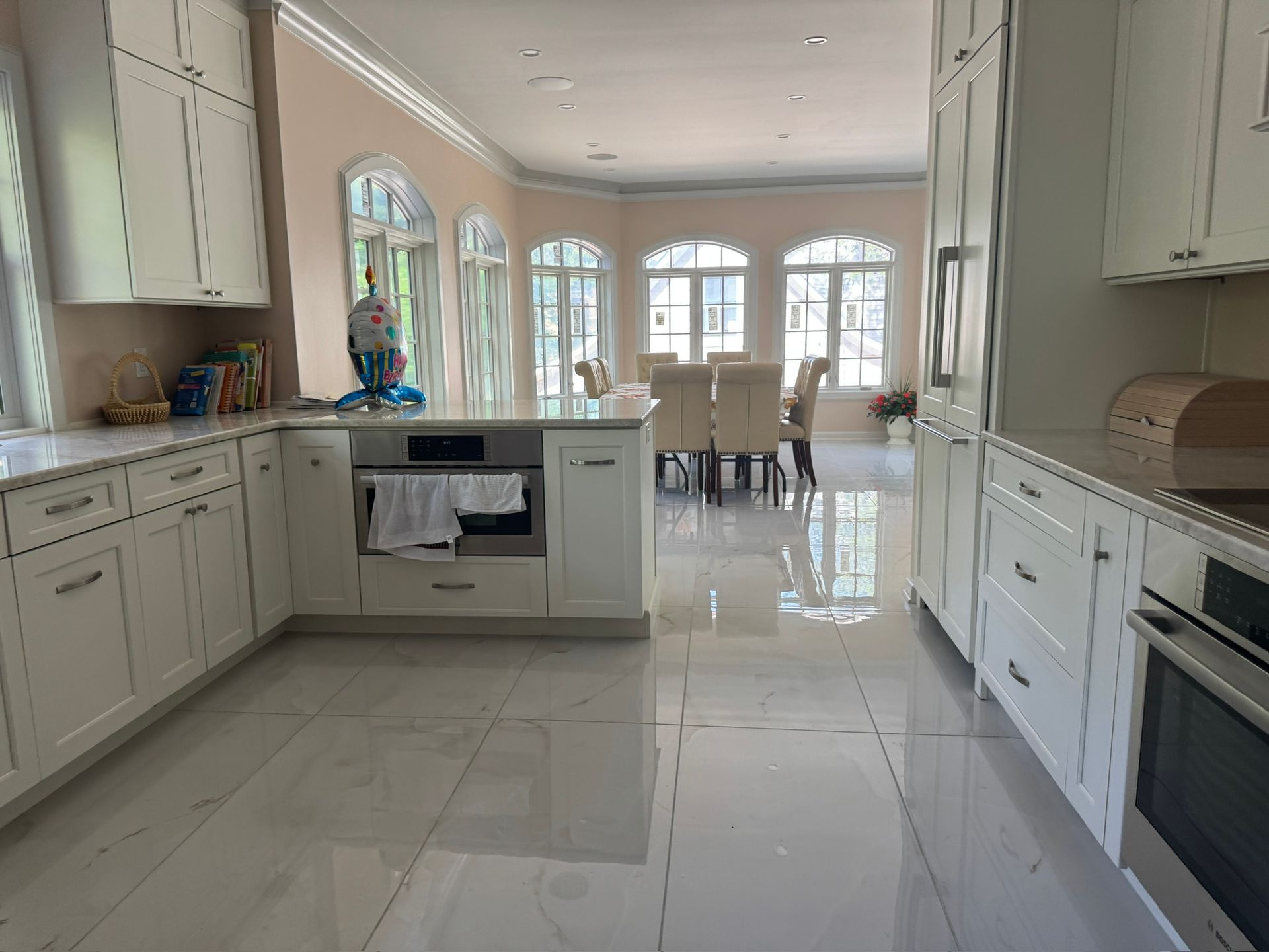 Bright white modern kitchen with glossy tile floor, cabinets, island, and arched windows in back.