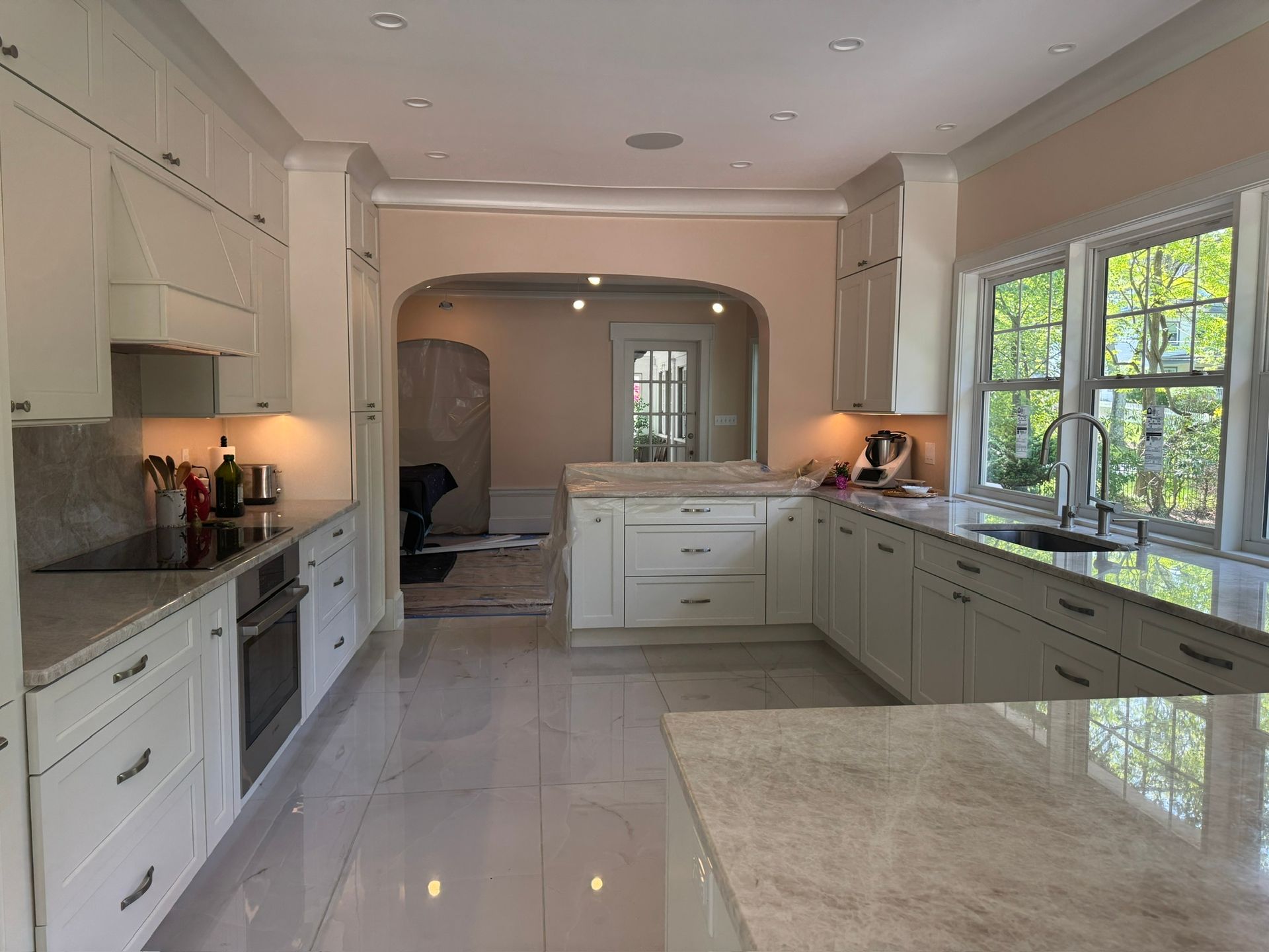 Bright white kitchen with long counters, tiled floor, and large windows overlooking greenery
