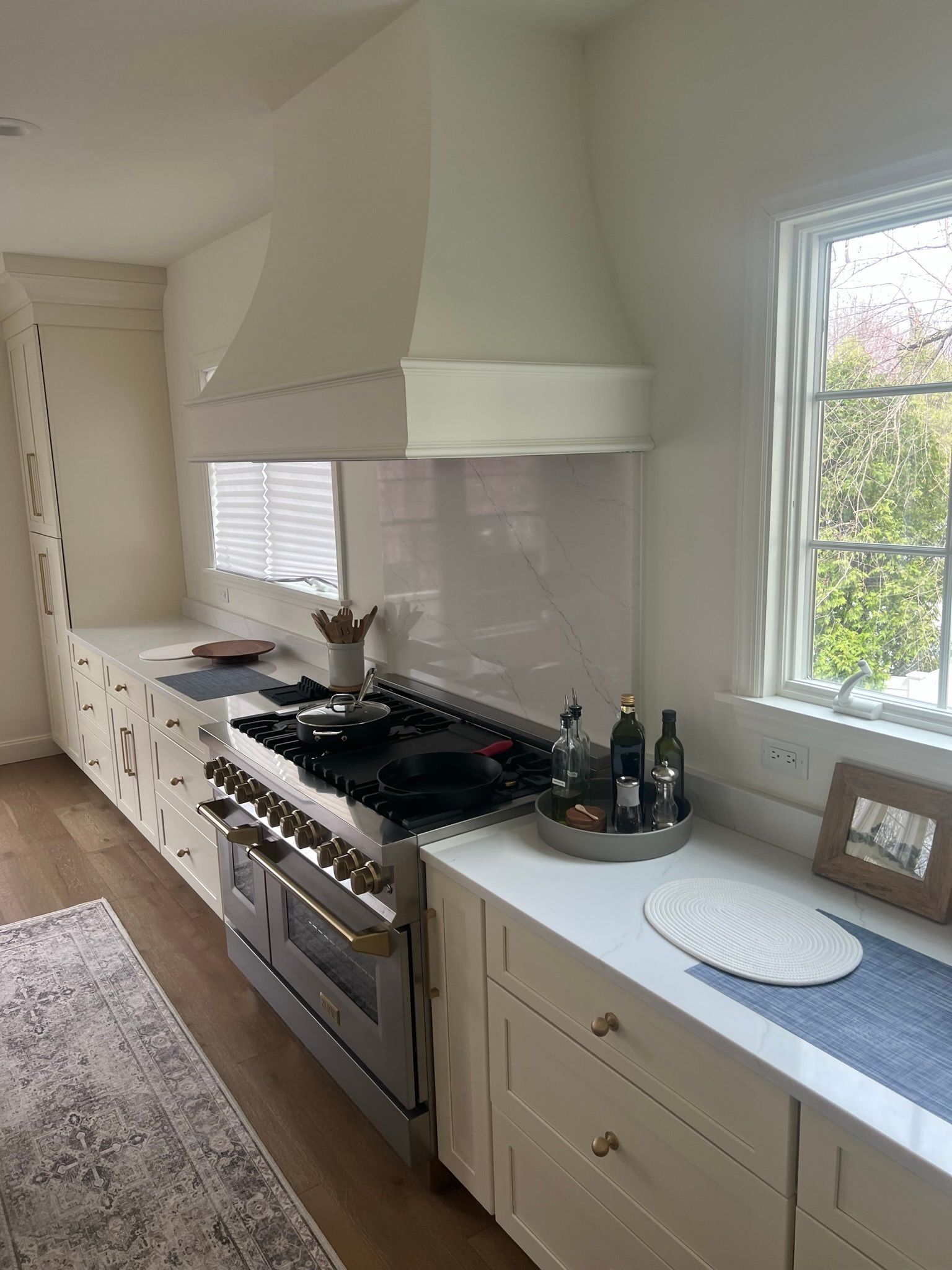 Bright white kitchen with long countertops, stainless stove, hood, and window over wood floors