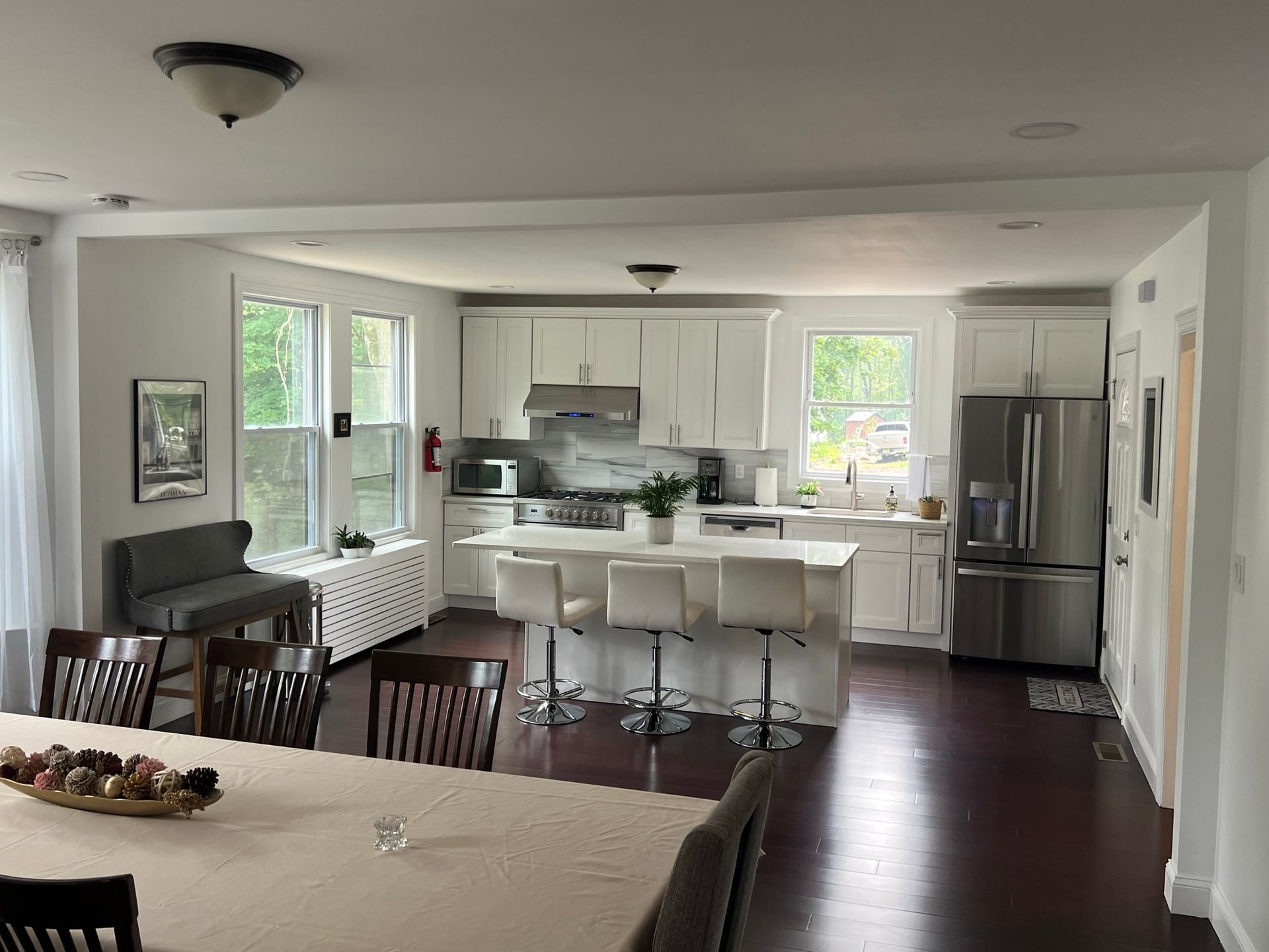 Bright open-plan kitchen and dining area with white cabinets, bar stools, dark wood floors, and large windows