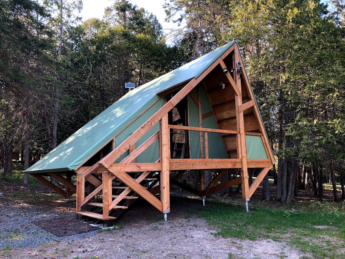 A-frame cabin made of wood with green roof in a forest setting; stairs lead up to a porch.