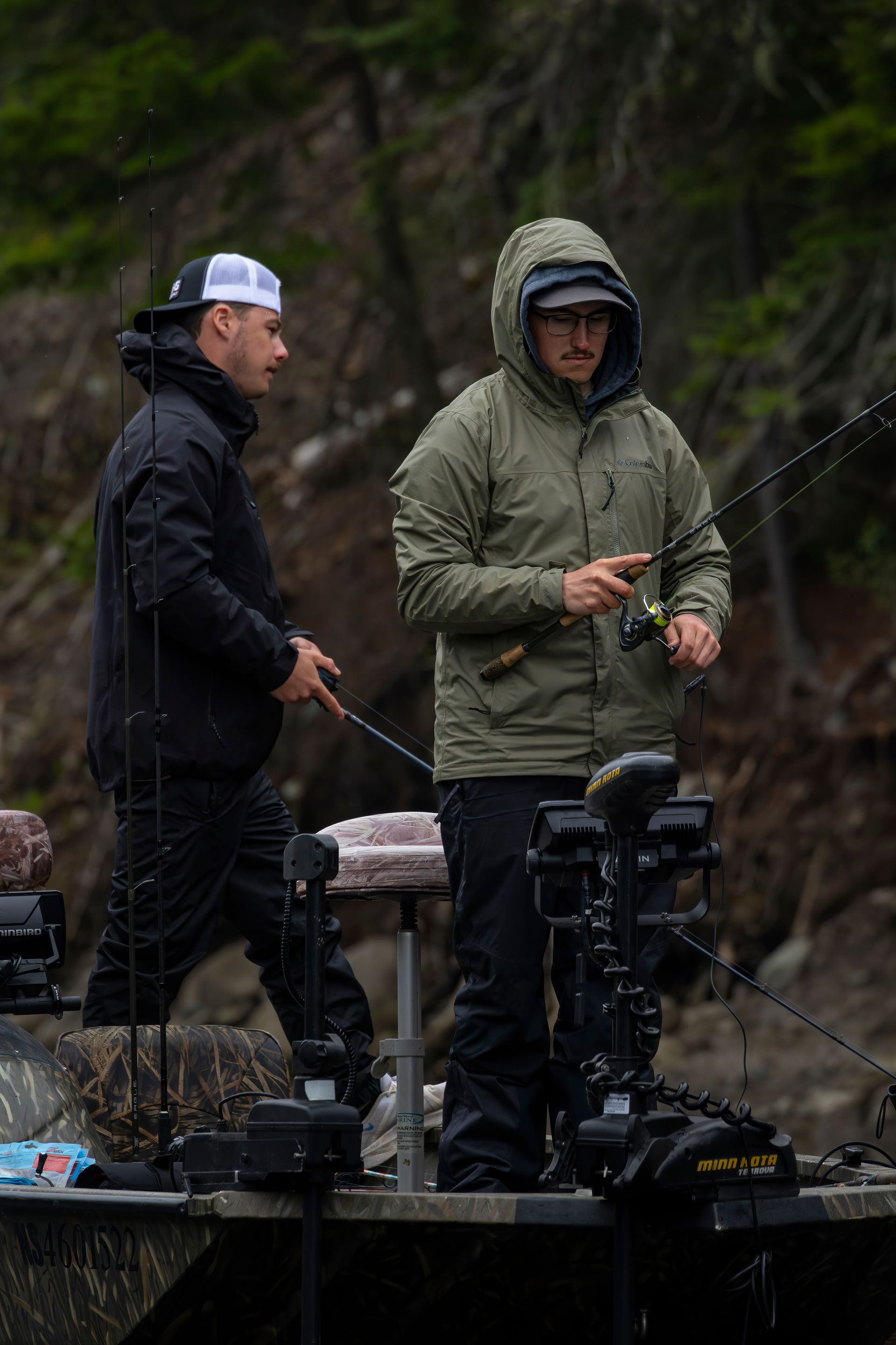 Two men fishing from a boat in a wooded area. One wears a black jacket and the other, a green hooded jacket.