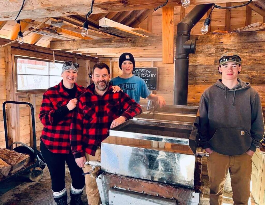 Family around a maple syrup evaporator in a rustic sugar shack. Red plaid shirts, steam rising.