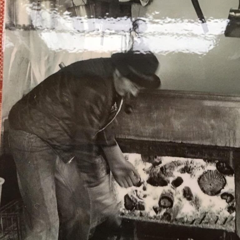 Man in hat, leaning, working on a dark surface; smoking pipe visible.