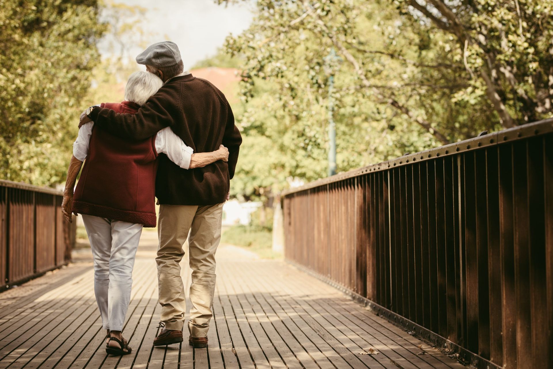 Elderly couple with arms around each other walking on a wooden bridge outdoors.