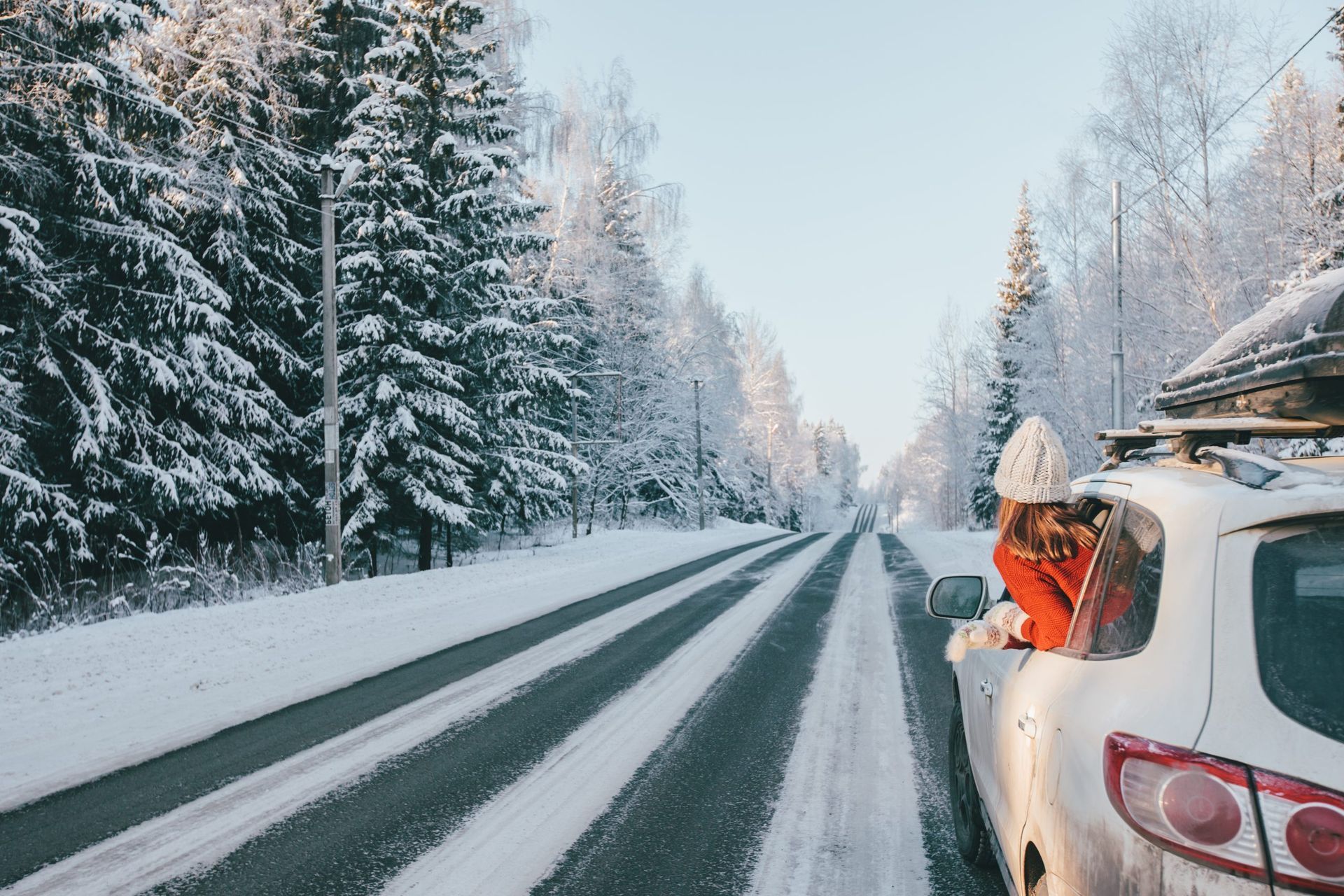 Person leans out of a white car on a snowy road, surrounded by snow-covered trees.