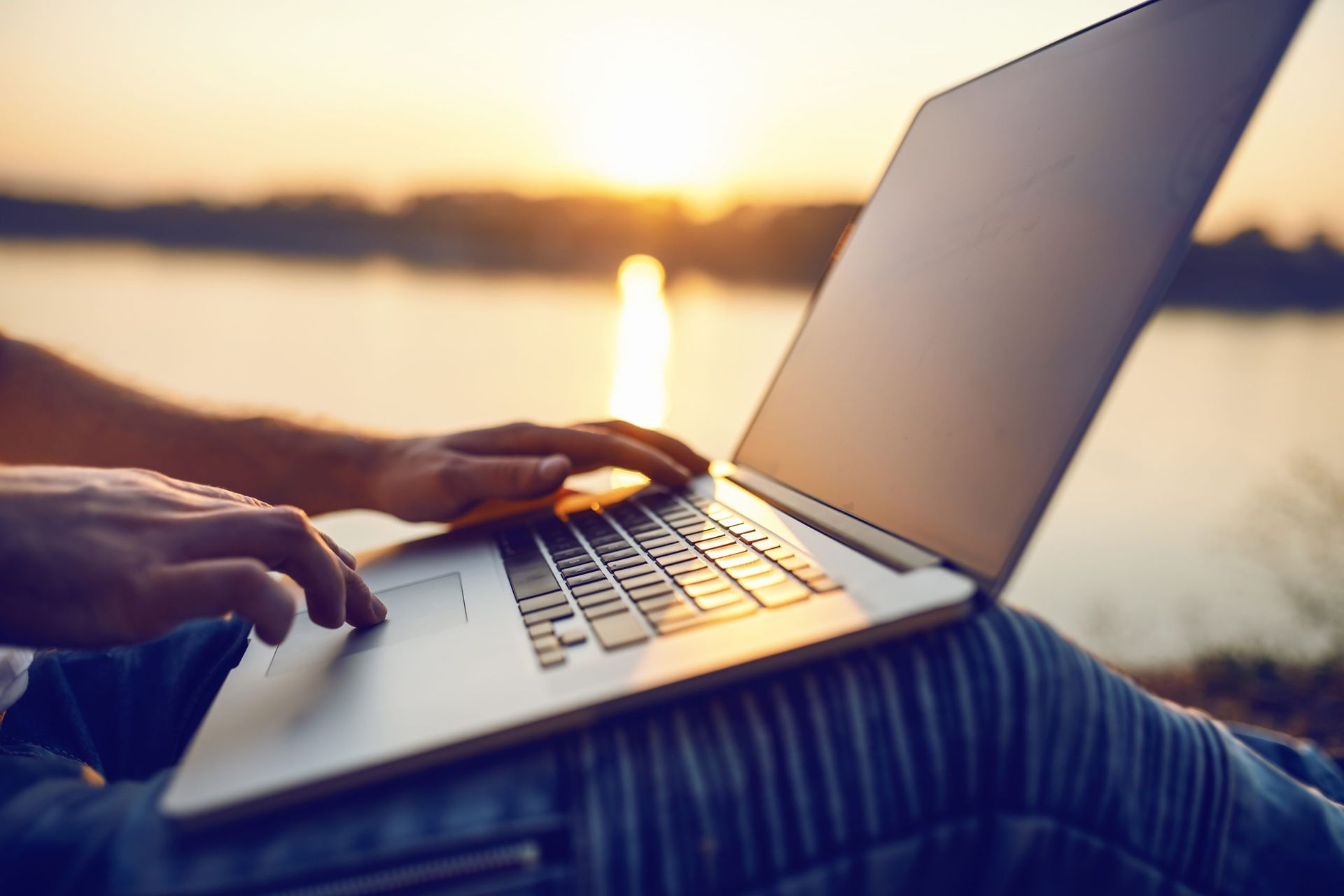 Person typing on a laptop outdoors near water with a sunset in the background.